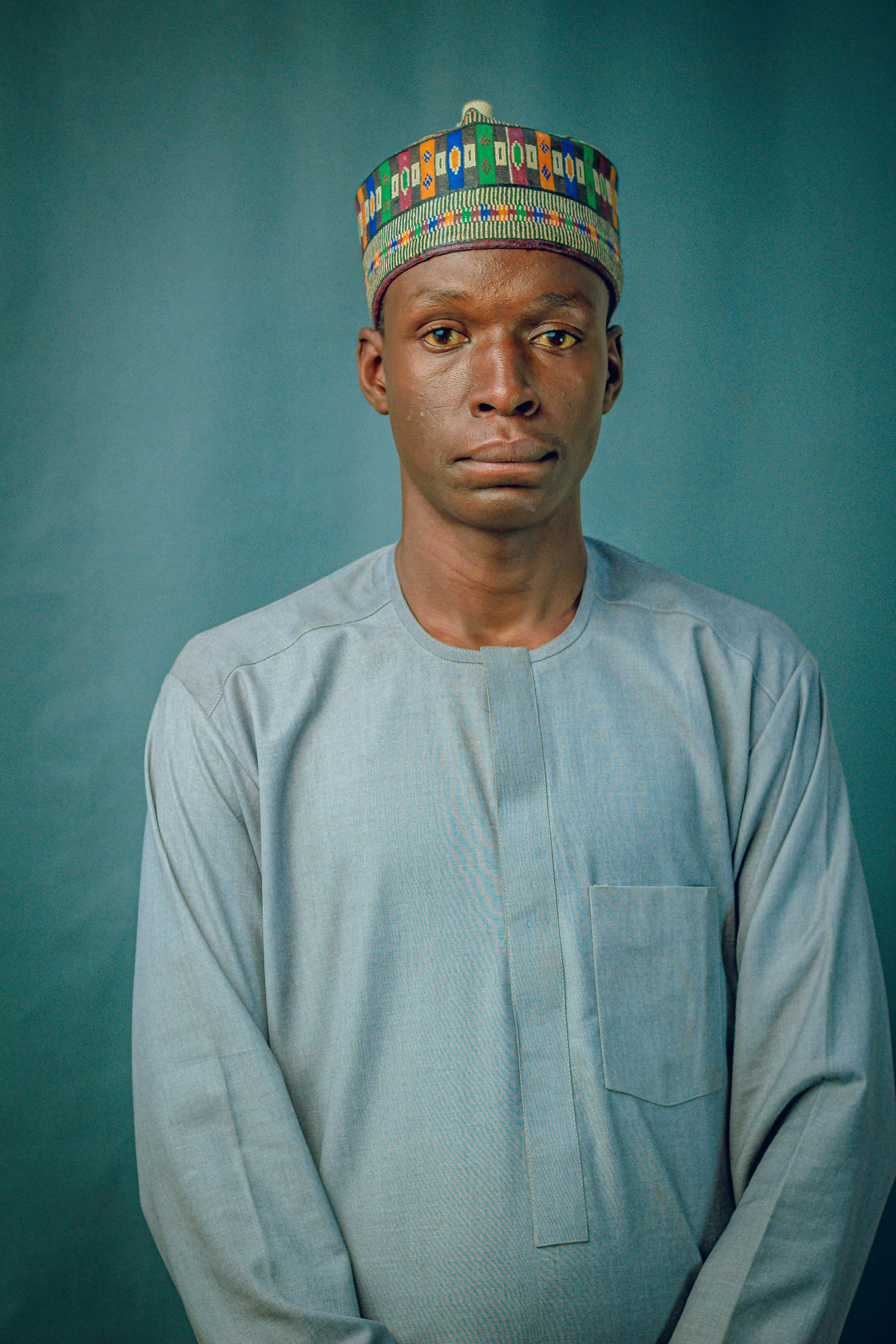 A solemn portrait of an adult man wearing traditional African clothing and a colorful cap.