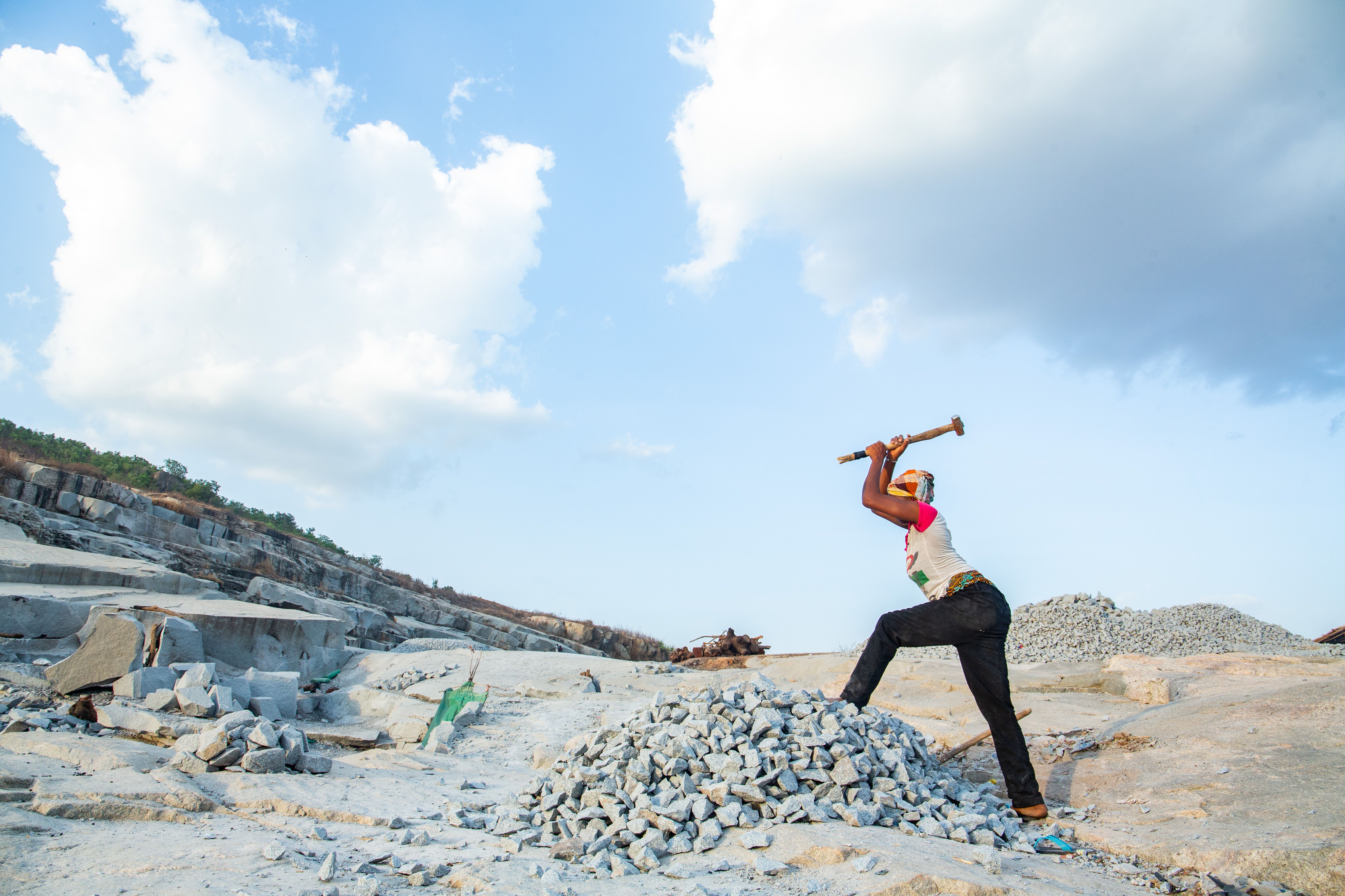A woman uses a hammer to break stones in an open quarry under a bright blue sky.