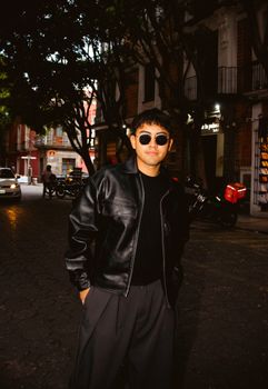 Man in a black leather jacket with sunglasses poses confidently on an evening city street.