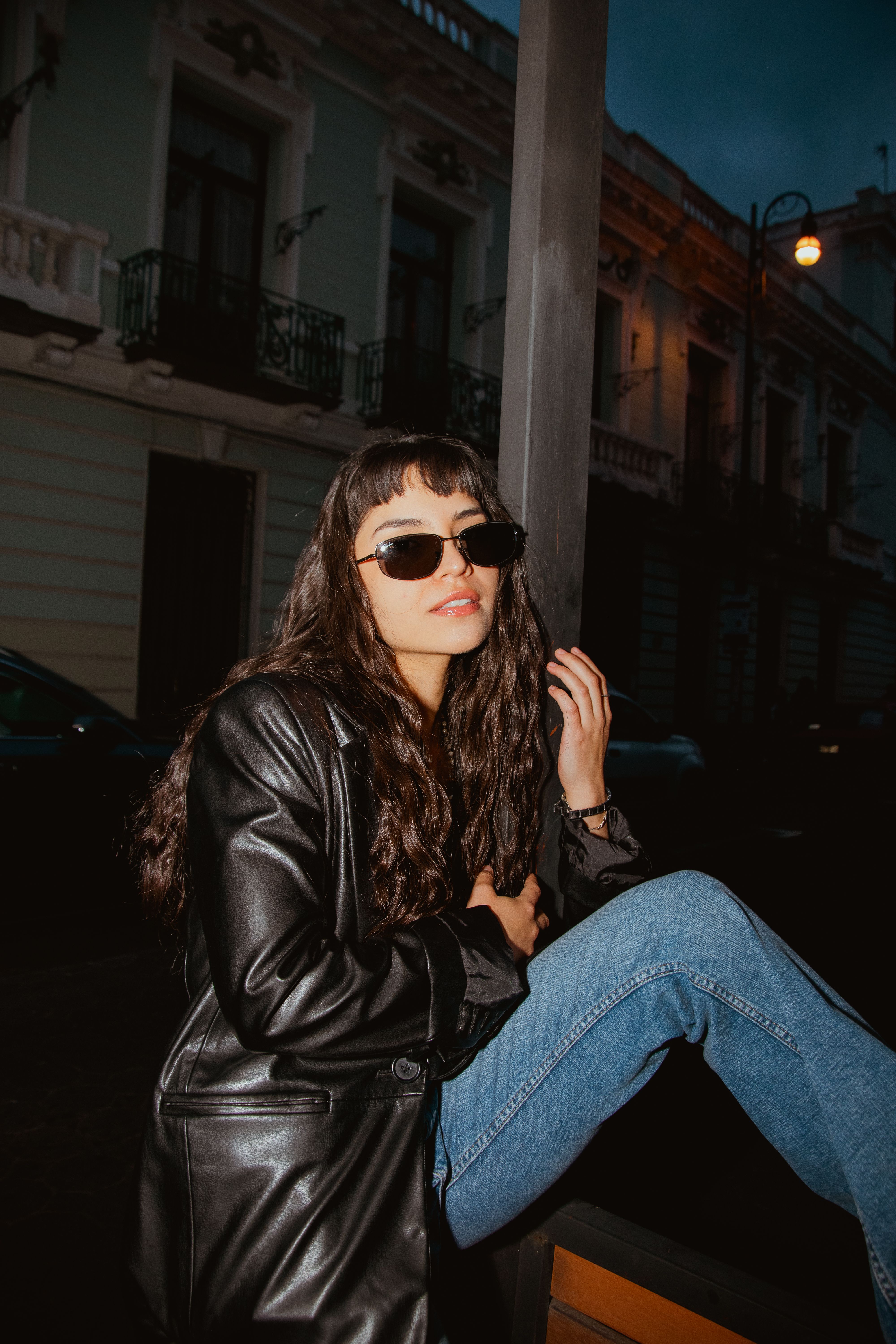 Fashionable woman wearing sunglasses and leather jacket in an urban night setting.