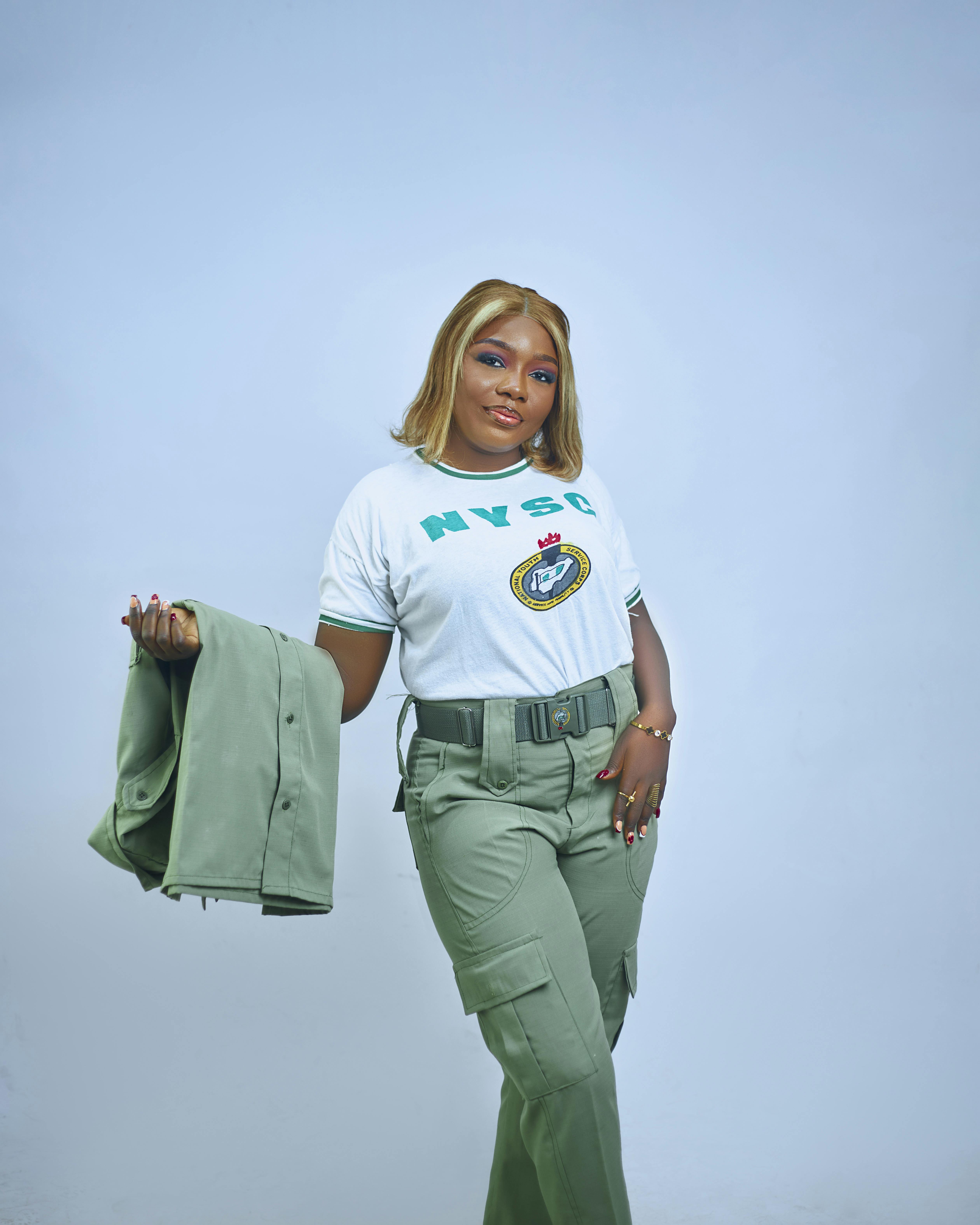 Smiling young woman poses confidently in NYSC uniform with blue backdrop.