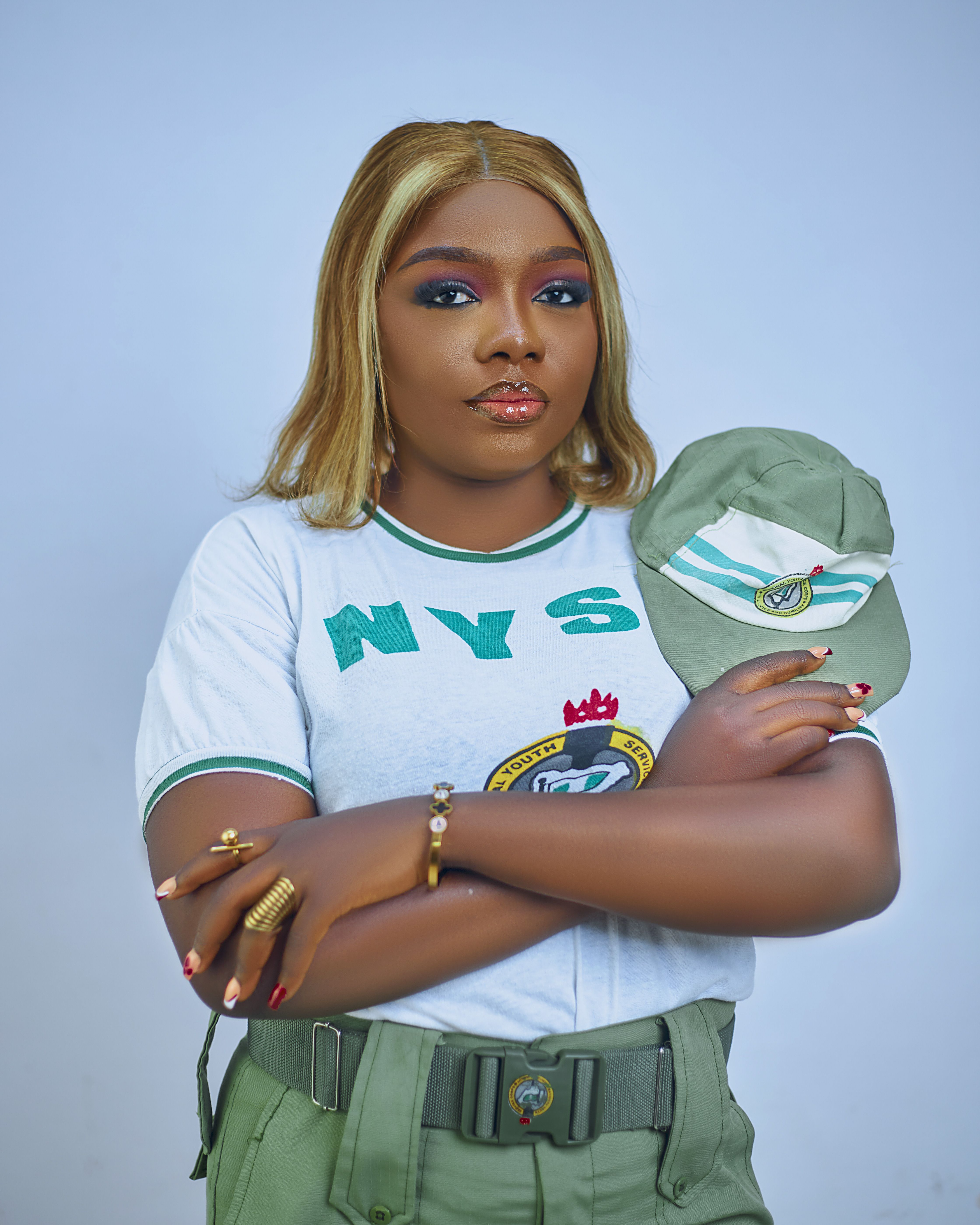 Portrait of a confident woman in NYSC uniform holding cap, white background.