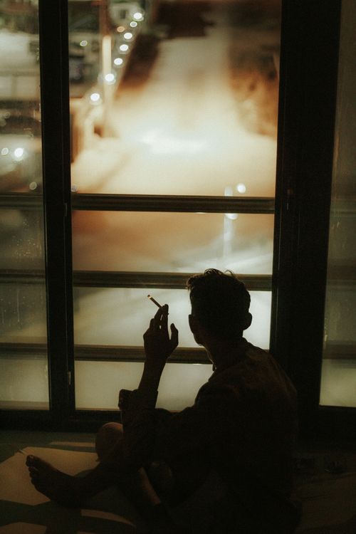 Free A thoughtful silhouette of a person smoking by a window, overlooking a nighttime scene. Stock Photo