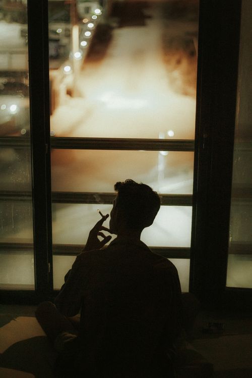Free A contemplative silhouette of a person smoking next to a foggy window overlooking a softly lit street at night. Stock Photo