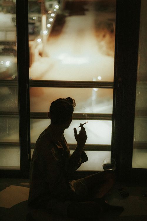 Free A contemplative silhouette of a person holding a cigarette by a window at night, capturing city lights. Stock Photo