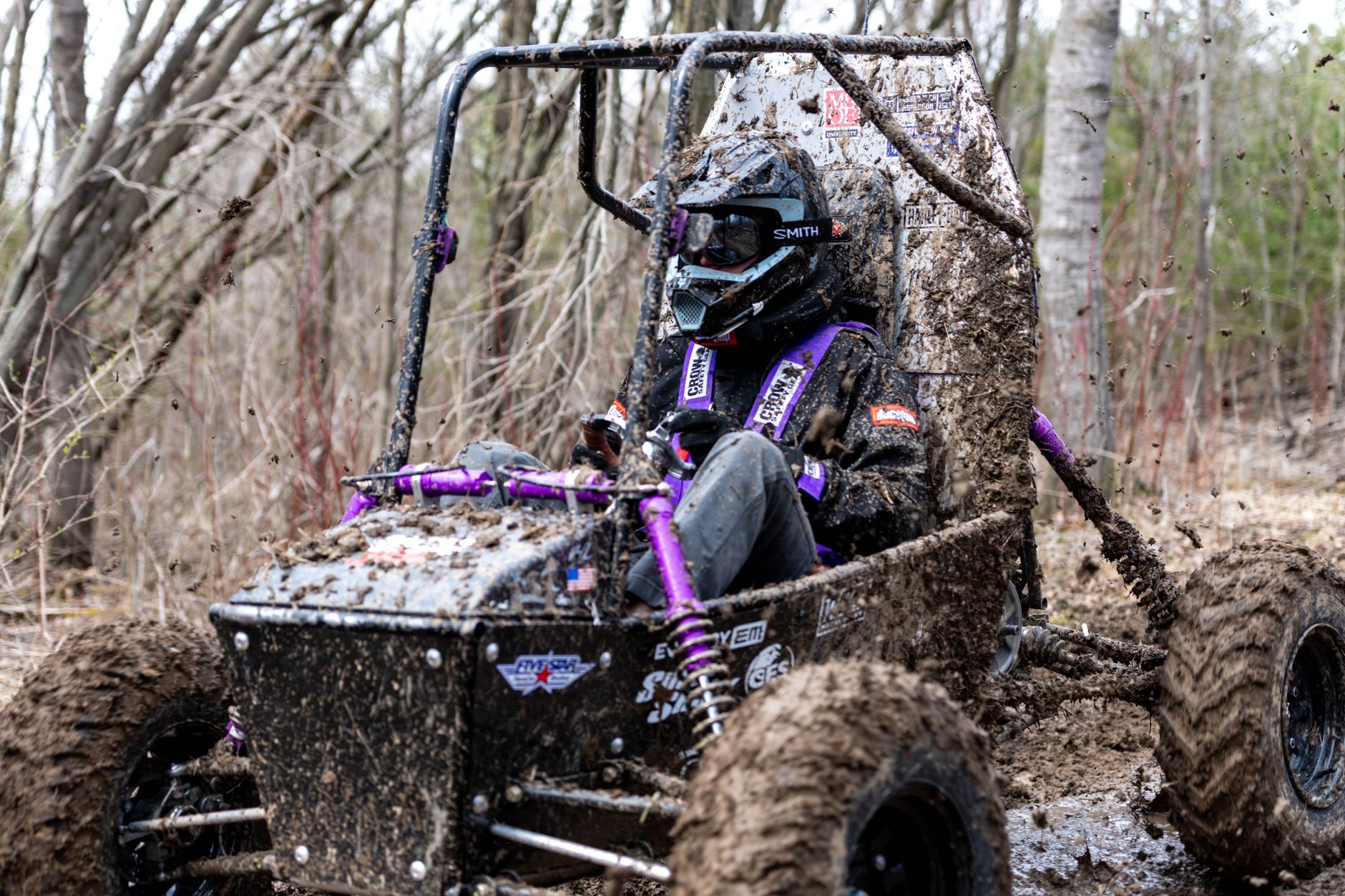 Driver in off-road vehicle speeds through muddy forest, showcasing off-road racing dynamics.
