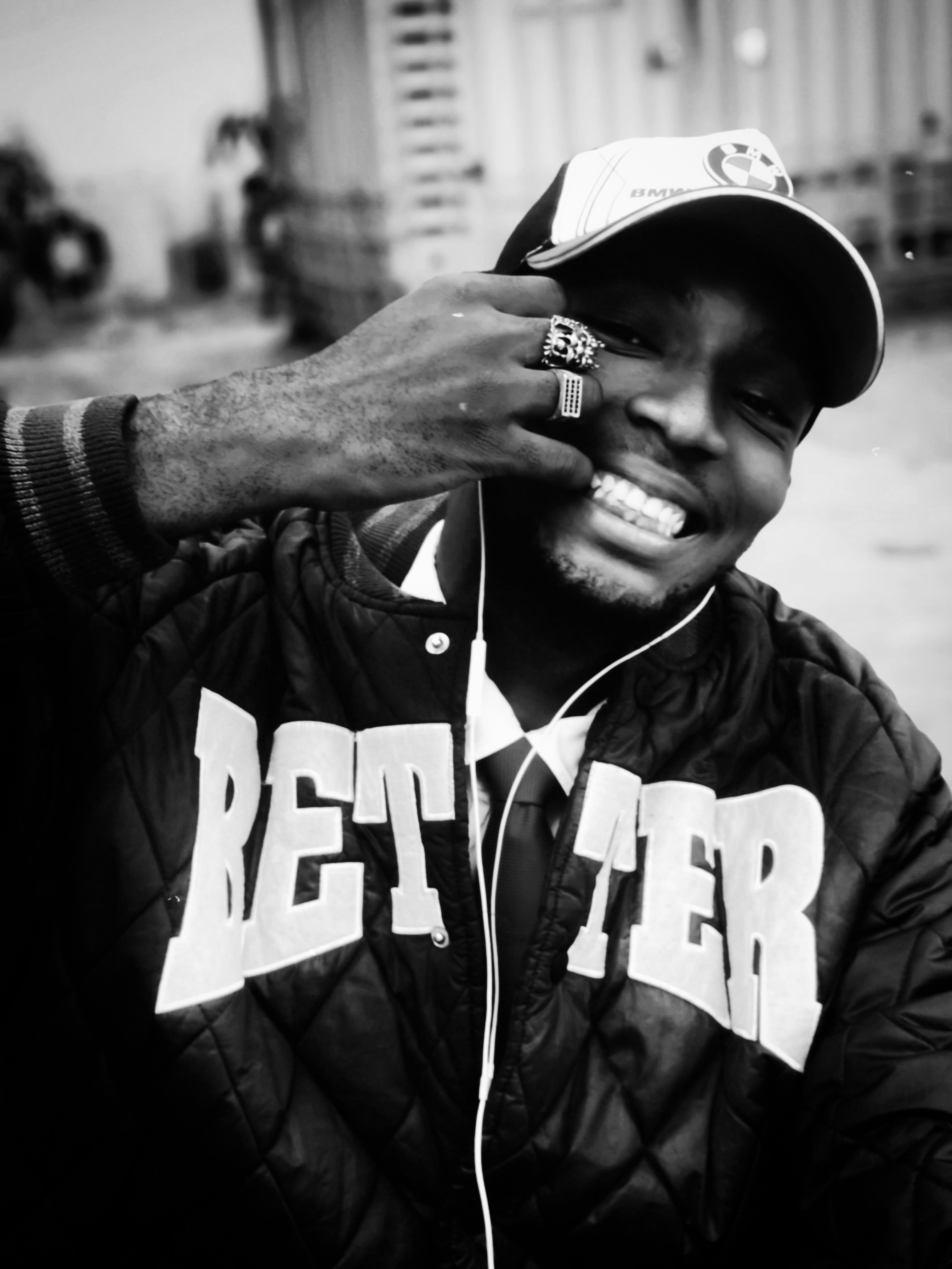 Black and white photo of a smiling man wearing a jacket and a cap, showcasing unique style.