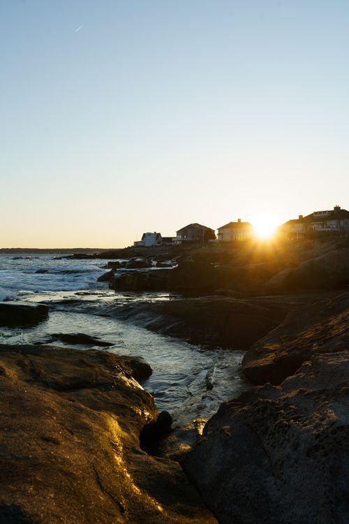 Free A beautiful sunset over rocky coastal shores with houses silhouetted against the sky. Stock Photo