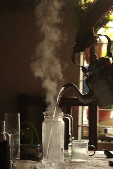 A steaming kettle pouring hot water into a glass jar in a cozy indoor setting.