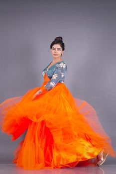 Stylish portrait of an Indian model wearing a flowing bright orange dress, photographed in a studio setting.
