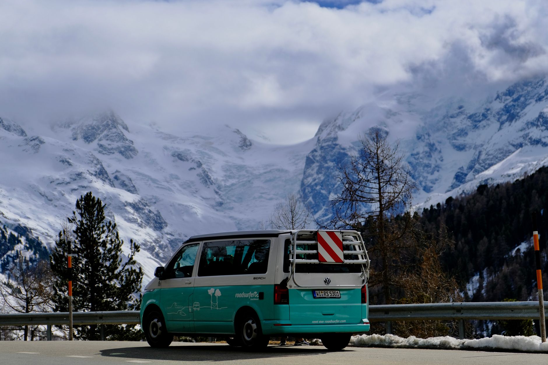 A scenic view of a teal van parked on a road with majestic snow-covered mountains in the background.