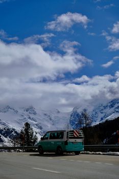 Turquoise van parked on a scenic mountain road with snow-capped alps in the background, under a clear blue sky.