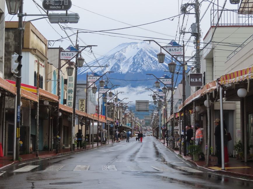 A picturesque Japanese street with Mount Fuji in the distance, featuring shops and pedestrians.