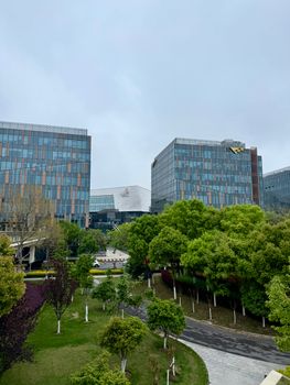 Contemporary skyscrapers with glass facades set against vibrant green trees under a cloudy sky.