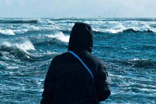 Free A person in a hooded black coat stands facing turbulent ocean waves under a cloudy sky. Stock Photo