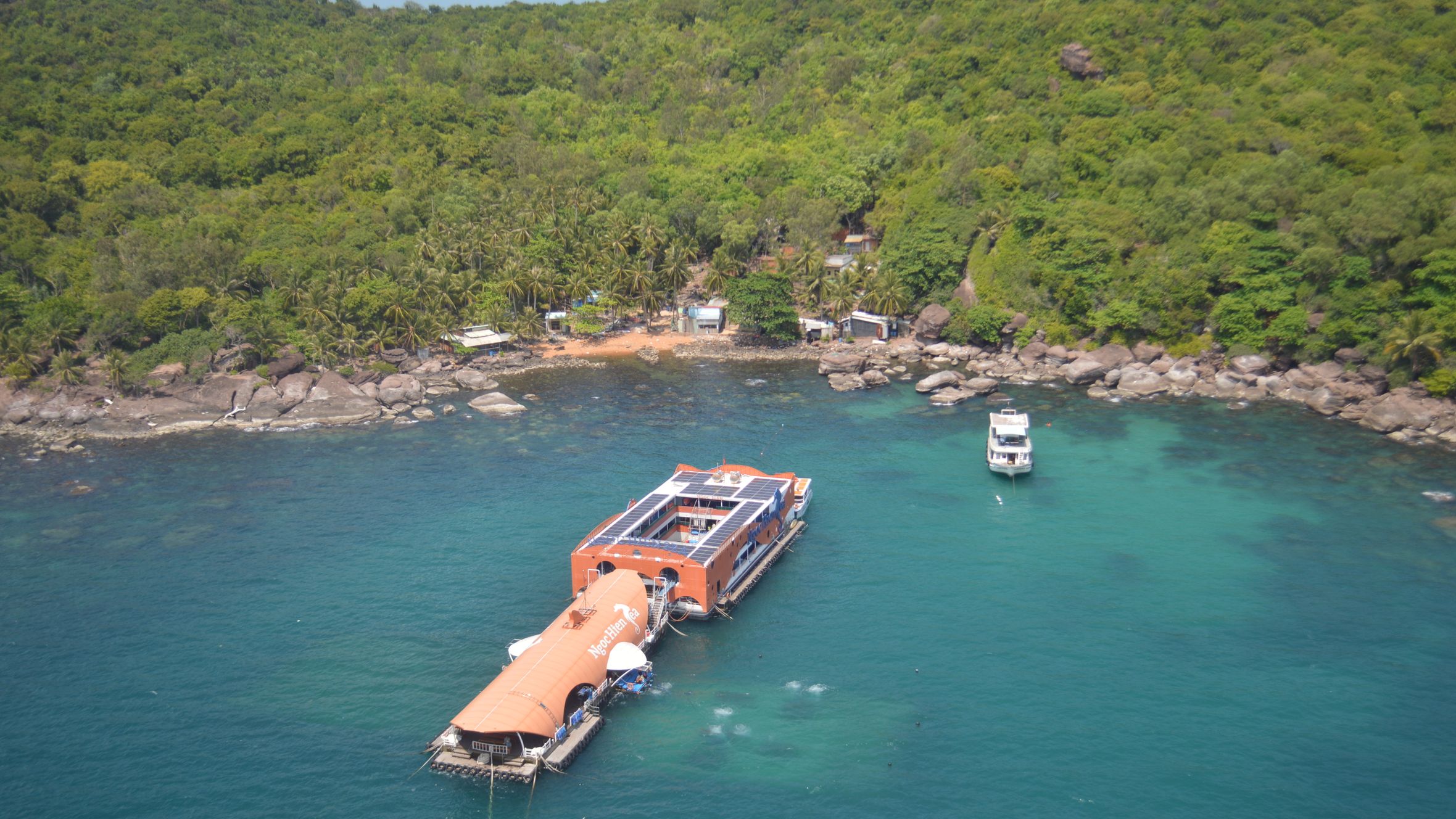 Aerial photo of a floating structure by a lush tropical island with boats in turquoise water.