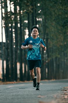 Asian male jogging on a tree-lined road, holding a bottle, showcasing dedication to fitness and healthy lifestyle.