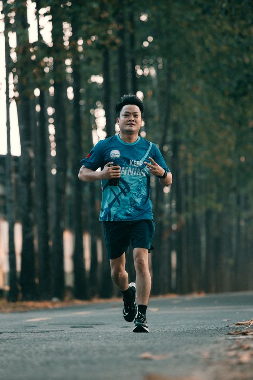 Free Asian male jogging on a tree-lined road, holding a bottle, showcasing dedication to fitness and healthy lifestyle. Stock Photo