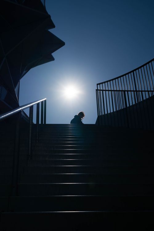 Free A lone figure in silhouette walks up a stairway under a bright sun, casting shadows. Stock Photo