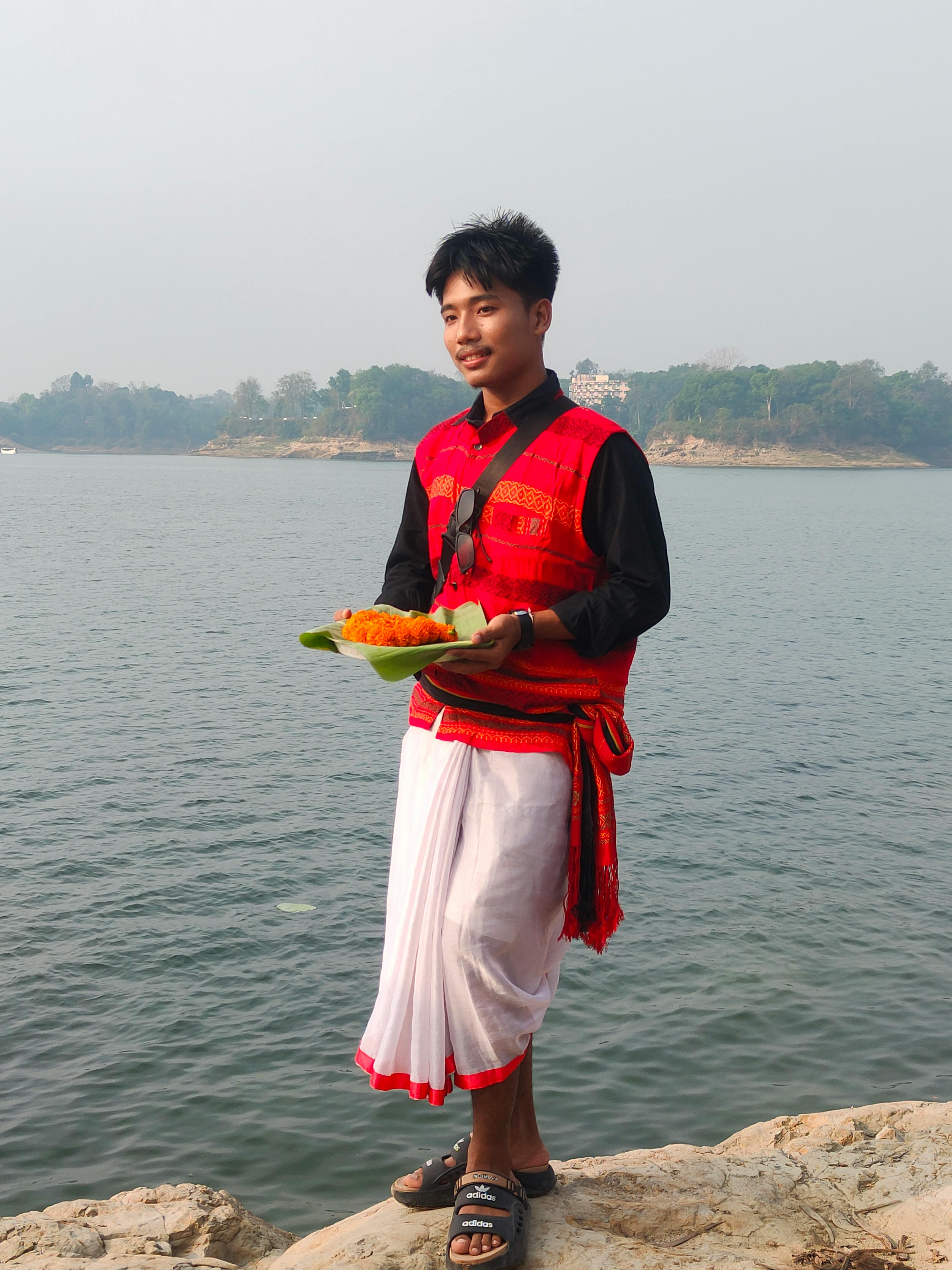 A man in traditional attire stands by a river holding a plate of food.