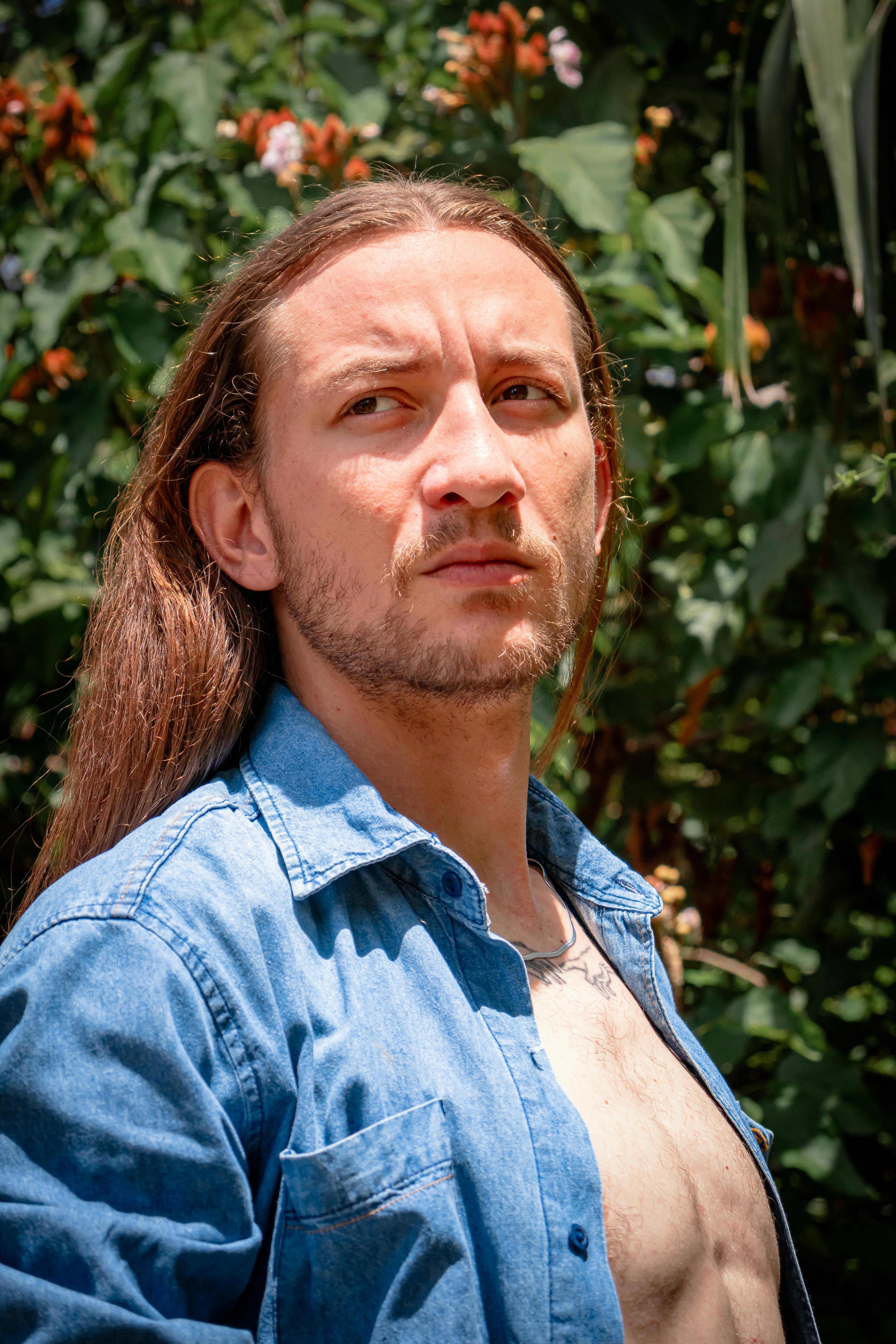 A man with long hair in a denim shirt looks thoughtful, surrounded by foliage.