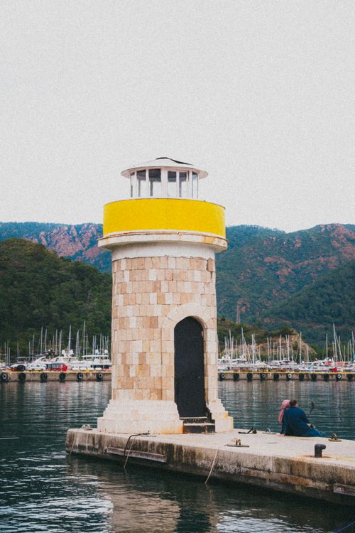 Free Photo of a tall white and yellow lighthouse beside a calm harbor with lush mountains. Stock Photo