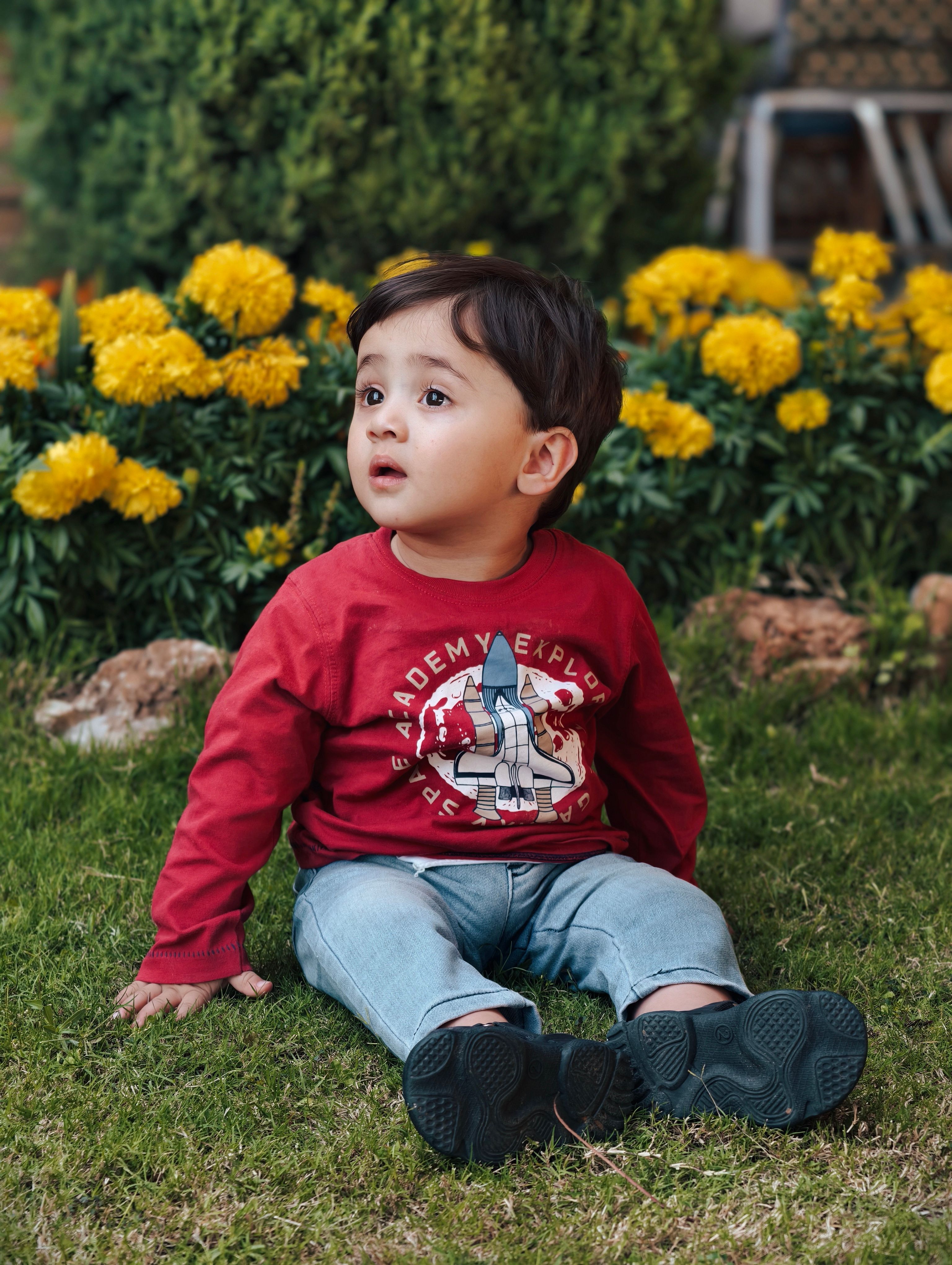 Adorable toddler in a red sweater sits on the grass with marigold flowers in the background.