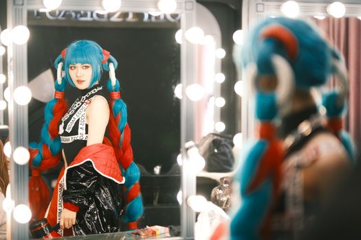Colorful cosplay character poses in a backstage dressing room with bright lights.
