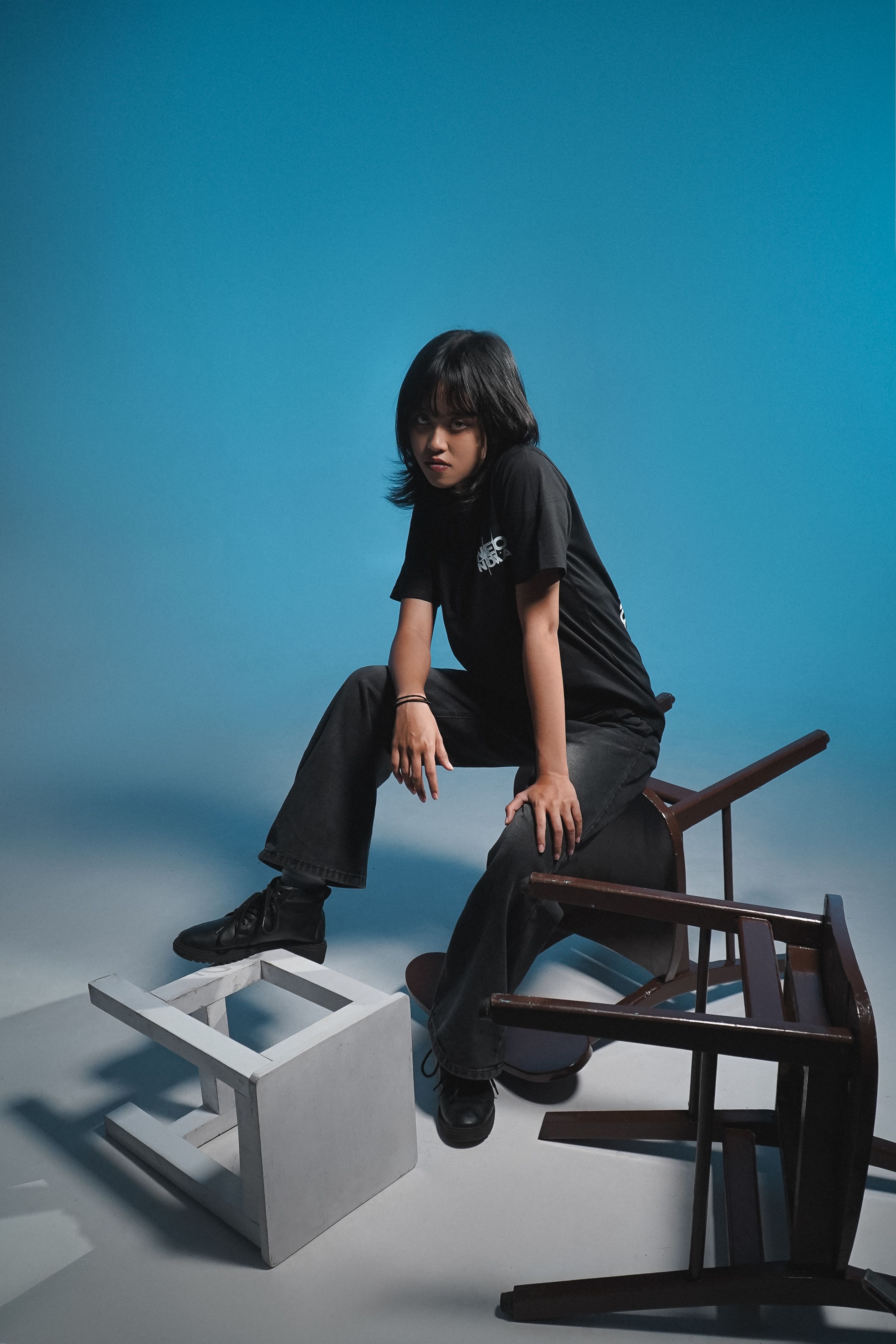 Teenage girl in a modern studio setting, posing with overturned chairs on a blue background.