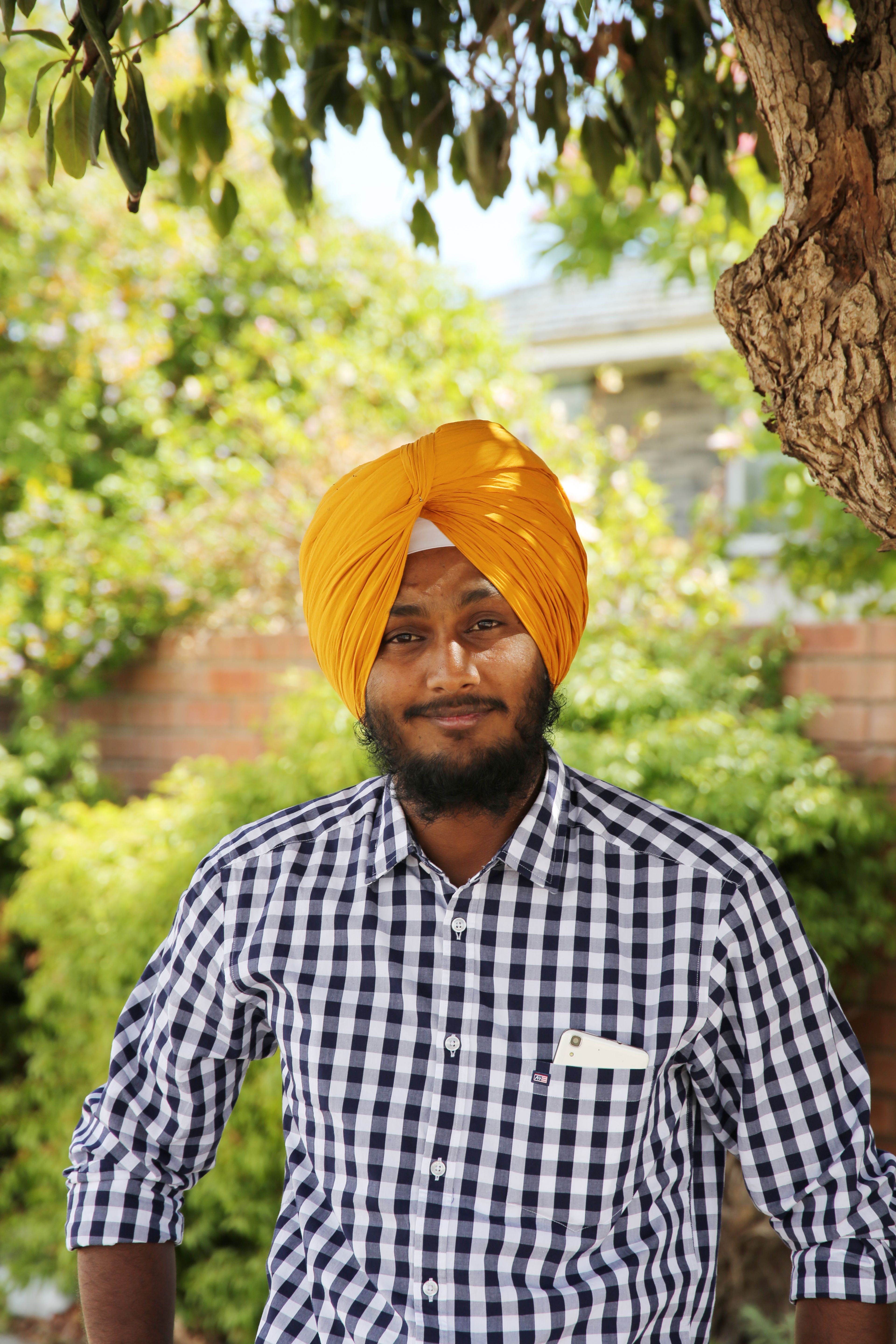 A man wearing an orange turban and checkered shirt smiles outdoors with greenery.