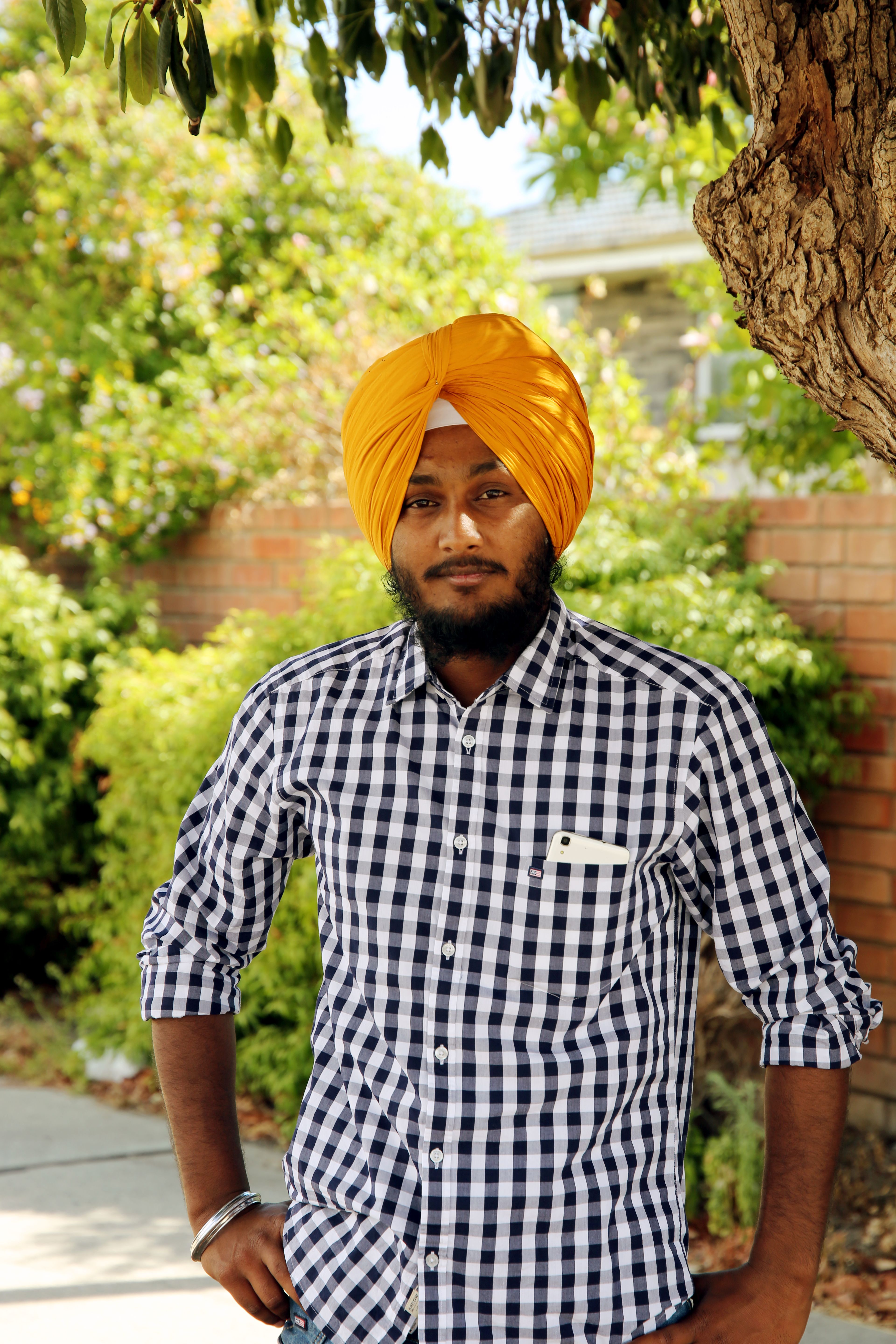 A confident man wearing a yellow turban and checkered shirt stands outdoors with a smile.