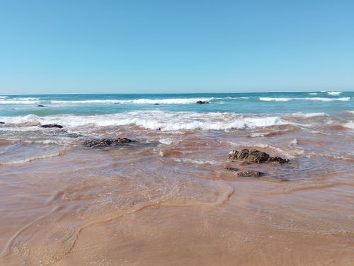 Free A peaceful beach scene with gentle waves lapping against exposed rocks, under a clear blue sky. Stock Photo