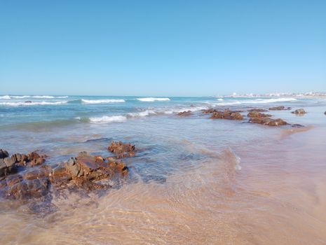 Tranquil beach view with gentle waves and rocky shoreline under a clear blue sky.