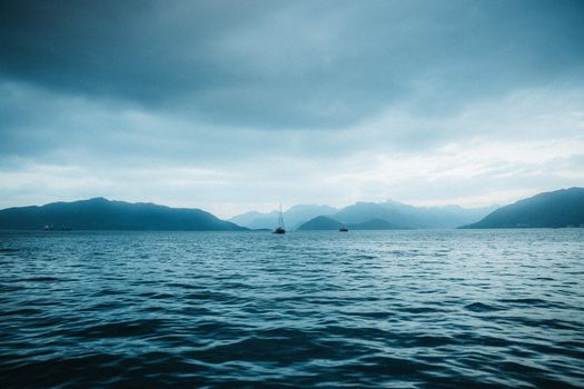 Calm ocean with distant sailboats under a moody sky and mountains in the background.