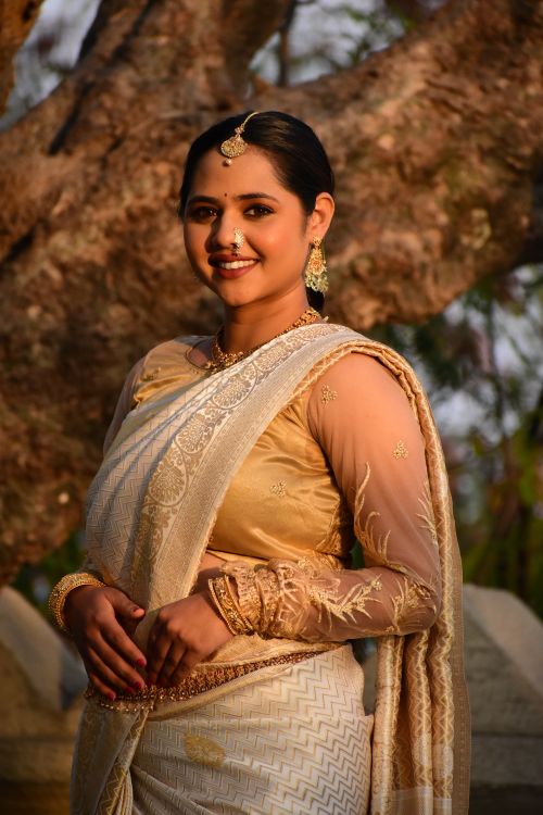 Free A woman in a beige saree adorned with traditional jewelry, standing outdoors with a tree backdrop. Stock Photo