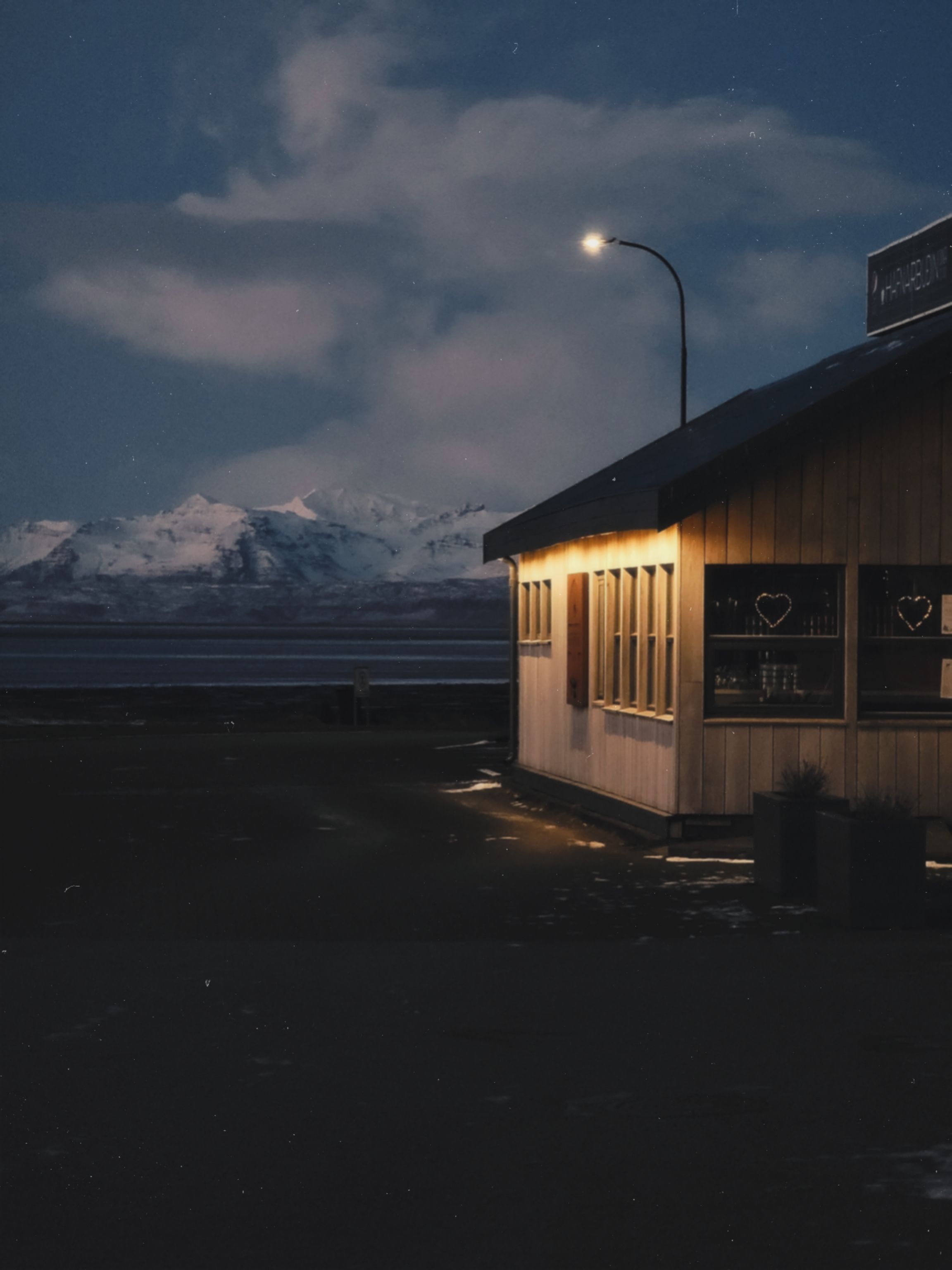Cozy roadside cabin glowing warmly against snowy mountains under a starlit night sky.