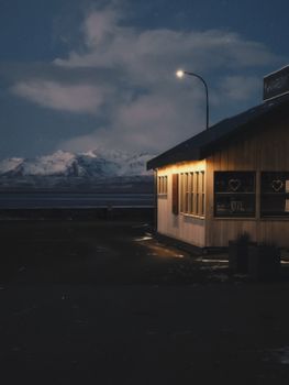 Cozy roadside cabin glowing warmly against snowy mountains under a starlit night sky.