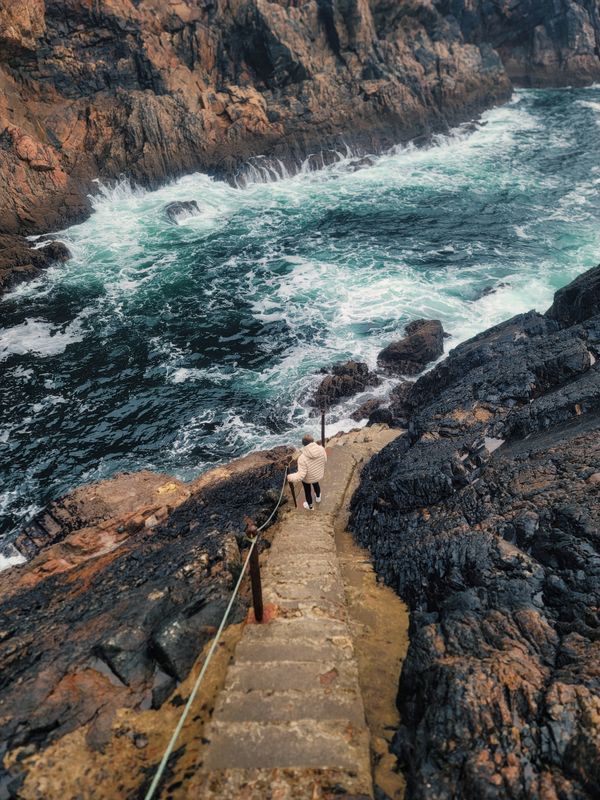 A person descends rocky steps by a rugged coast, amid crashing waves.