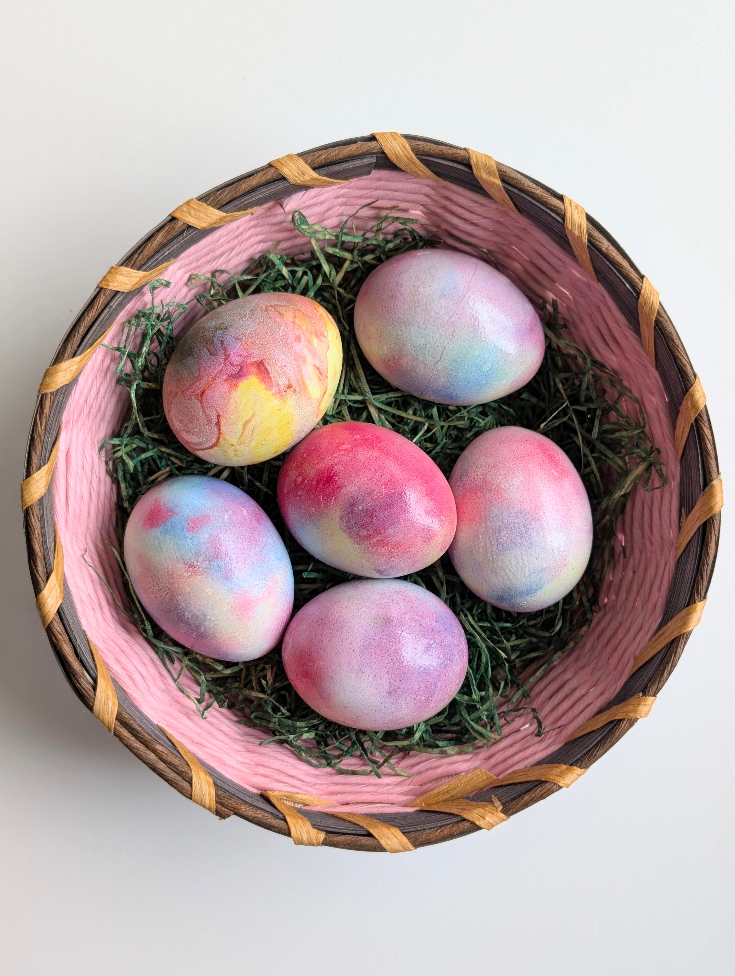 A dozen hard-boiled eggs dyed in pastel blue, pink, yellow, and purple, drying on a wire rack next to a bowl of dye cups.