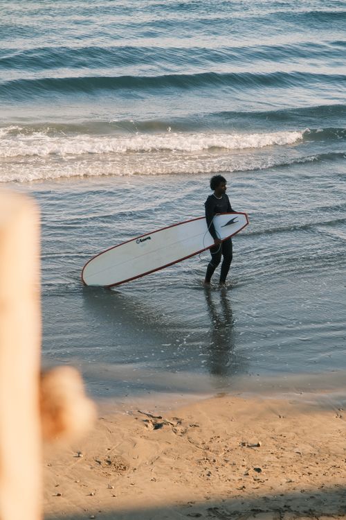 Free Surfer holding a long surfboard while walking along a serene beach shore. Stock Photo