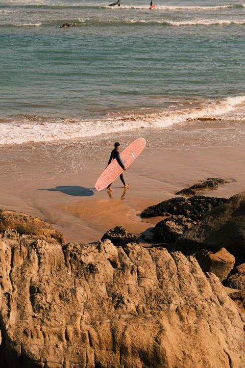 Free A lone surfer carries a pink surfboard along a rocky beach at sunset, embracing seaside adventure. Stock Photo