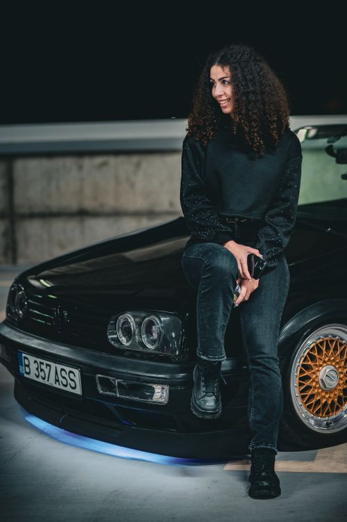 Free Casual portrait of a young woman seated on a stylish vintage car in a dimly lit setting. Stock Photo
