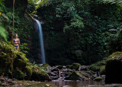 Woman in bikini by a serene jungle waterfall, surrounded by lush greenery and rocky terrain.