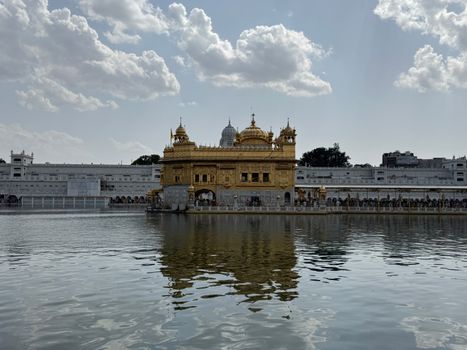 Stunning view of the Golden Temple reflecting in water under a bright, cloudy sky in Amritsar, India.