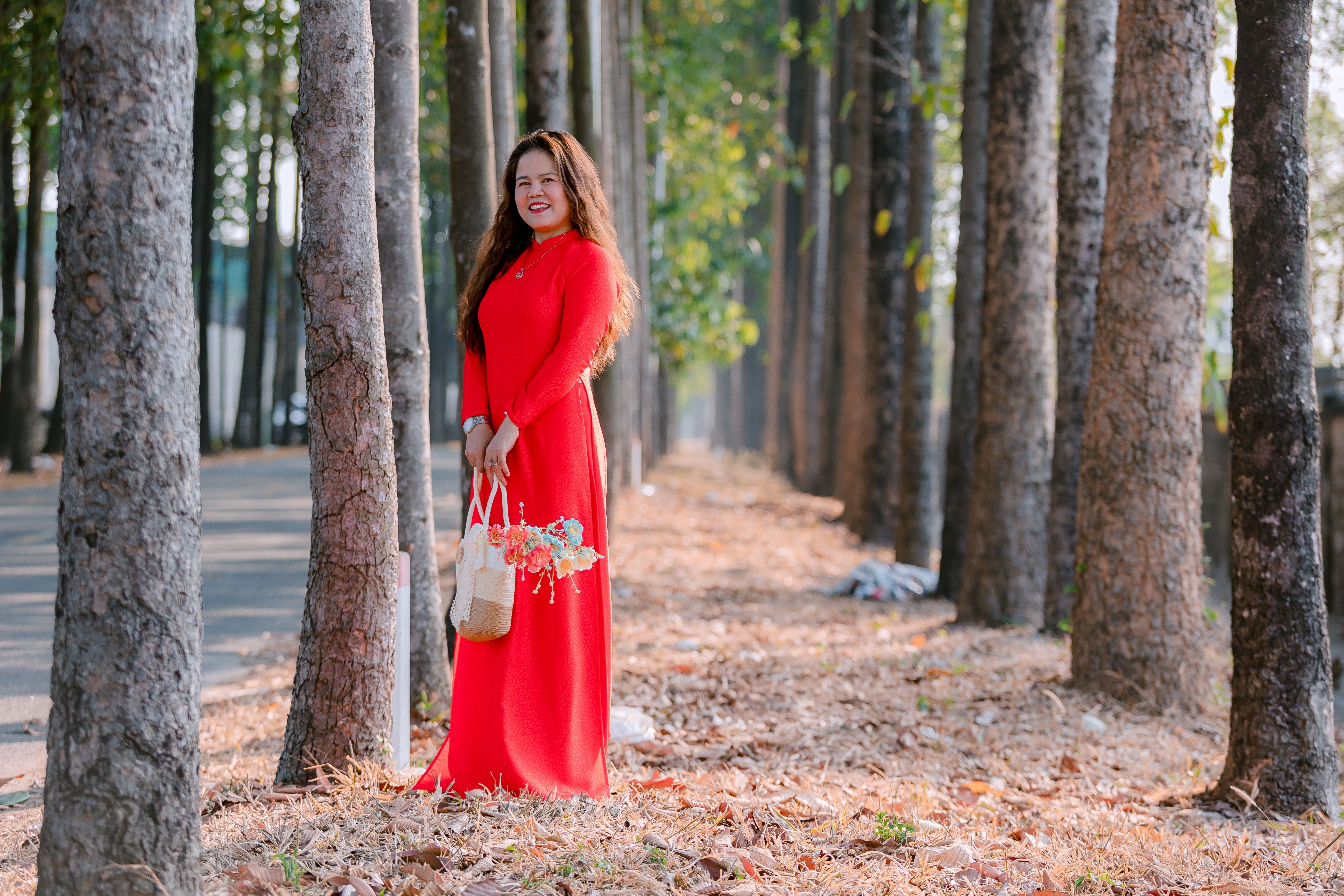 A woman in a vibrant red dress stands gracefully among tall trees, holding a flower bouquet.