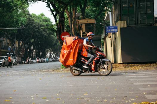 A delivery driver transports goods on a motorcycle in a bustling city street scene.