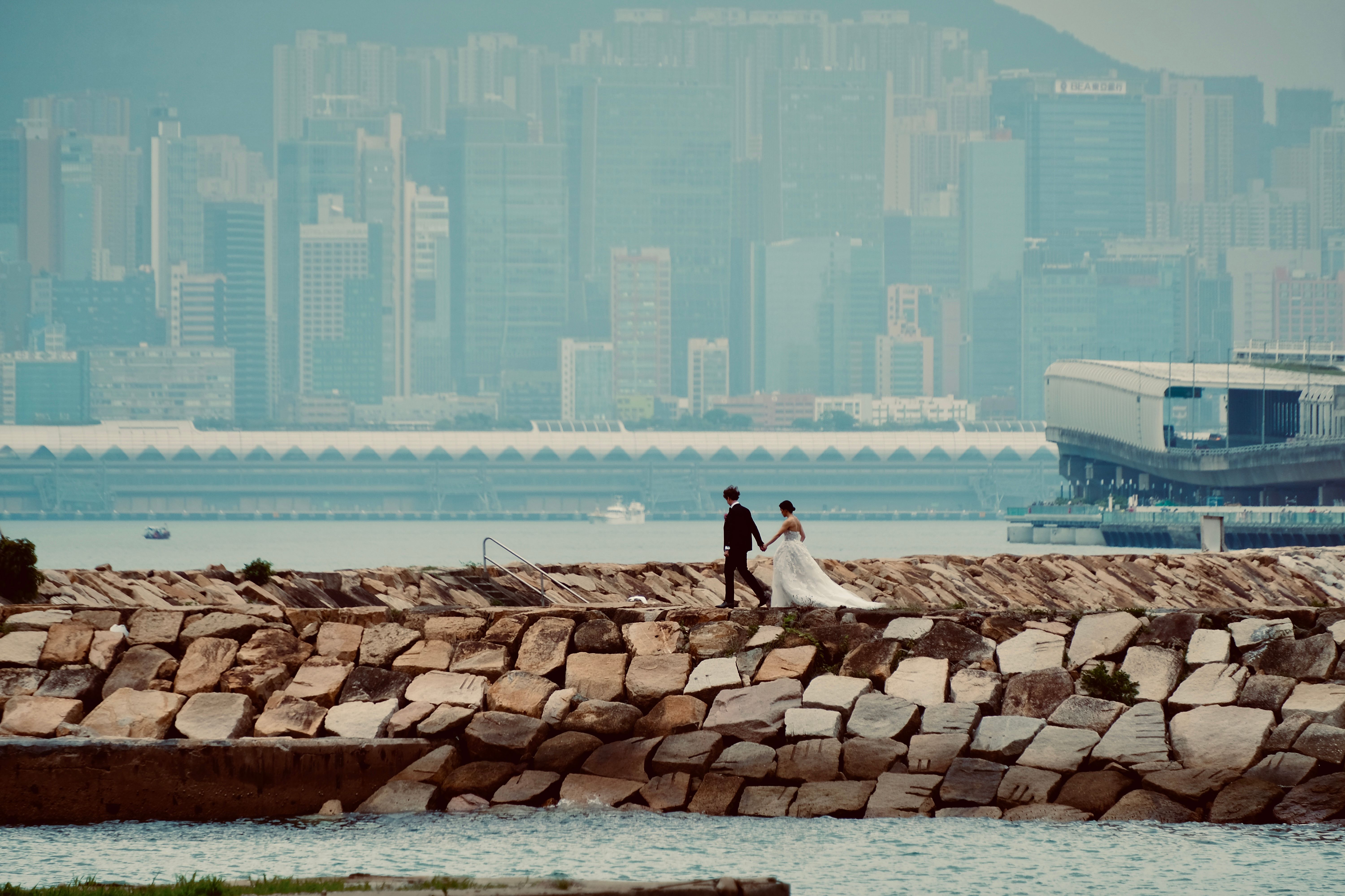A couple in wedding attire walks along the waterfront with Hong Kong skyline in the background.