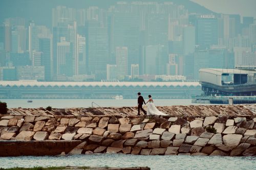 Free A couple in wedding attire walks along the waterfront with Hong Kong skyline in the background. Stock Photo