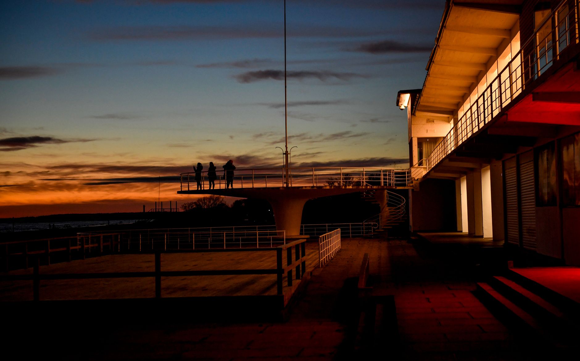 Silhouettes on a seaside platform at sunset in Pärnu, Estonia, featuring dramatic skies and illuminated buildings.