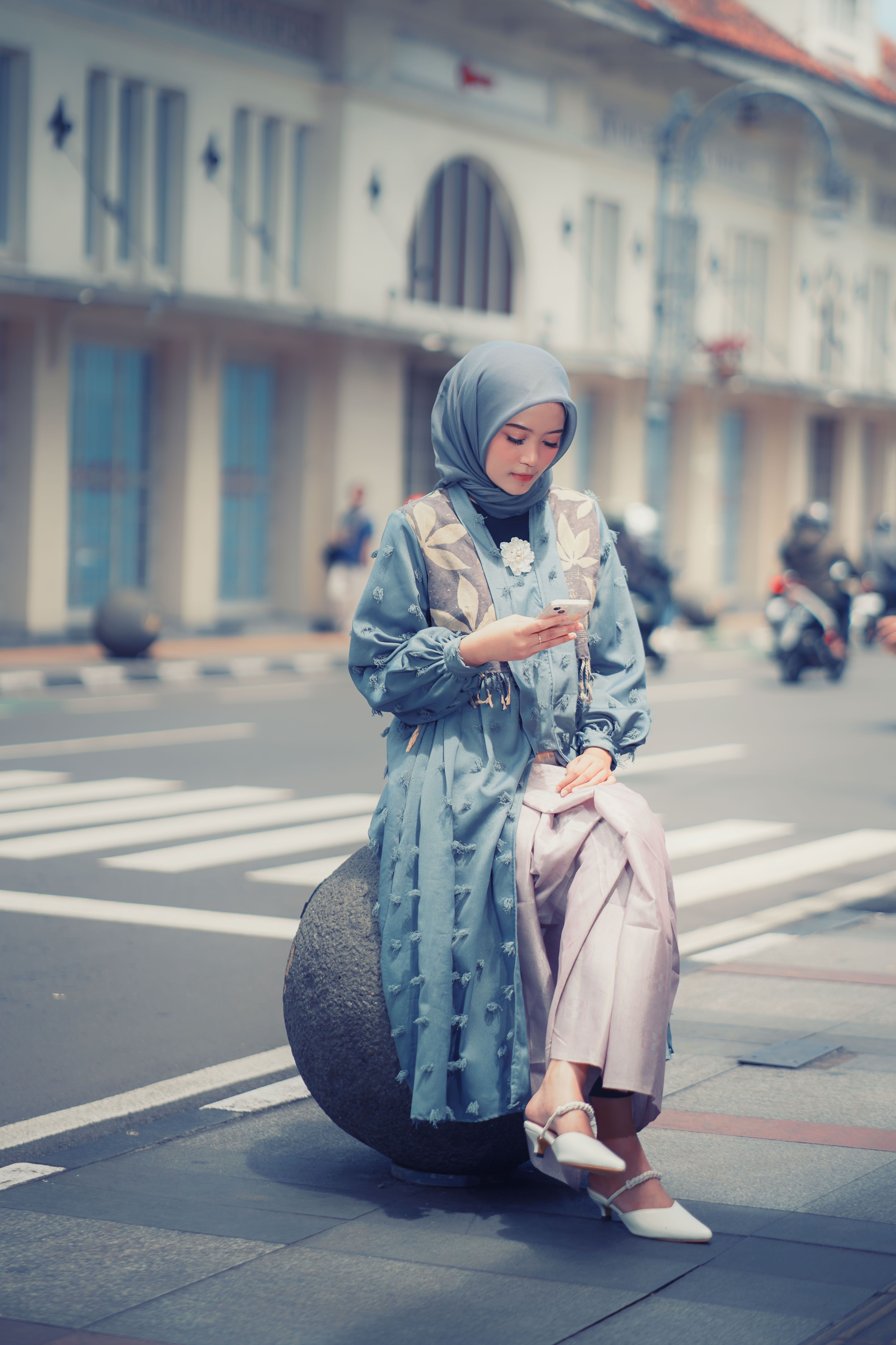 A woman in a blue hijab uses her phone while sitting on a city street in Bandung, Indonesia.